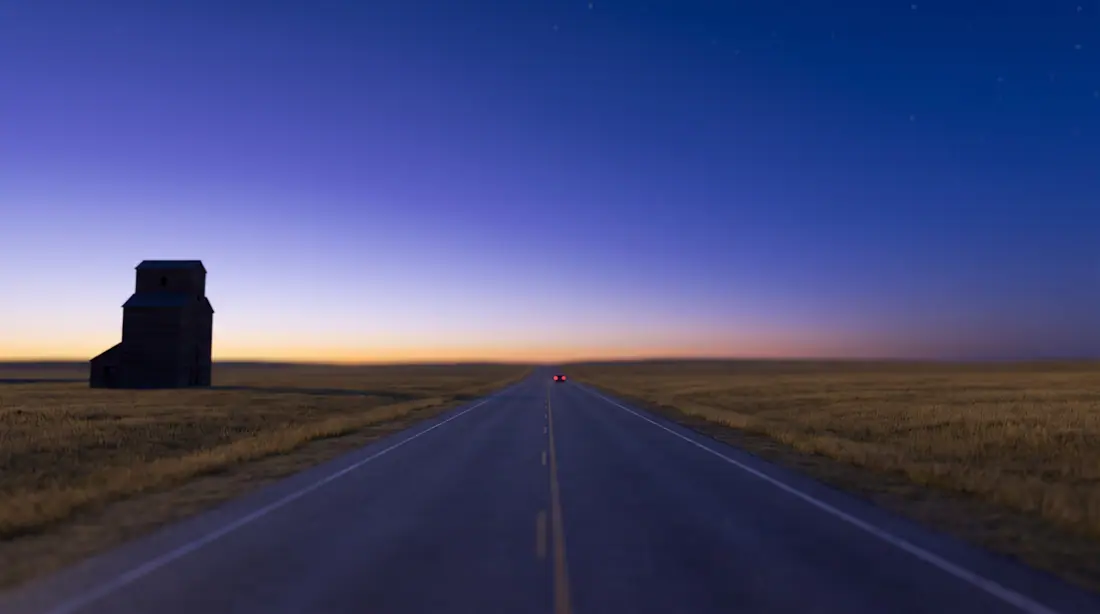 Straight two-lane prairie highway running to the horizon at blue hour, a lone grain elevator to one side