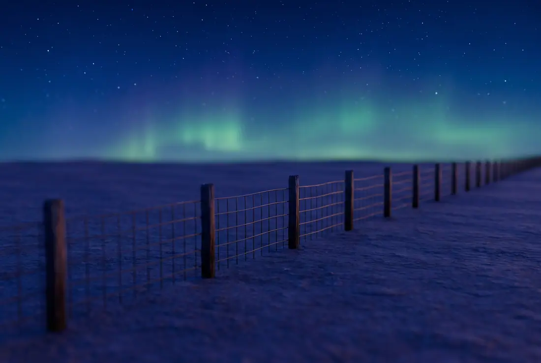 Star-filled night sky with a faint green aurora over flat prairie and a fence line